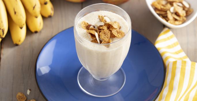 Imagem de um copo com a bebida cremosa em tom claro, decorado com cereal. Ele está sobre um prato azul e, ao redor, há cereais espalhados na mesa, uma penca de bananas, duas maçãs e um tecido listrado amarelo e branco.