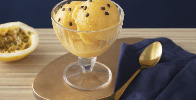 Foto em tons de amarelo da receita de sorbet de damasco e maracujá servida em uma taça de vidro sobre uma mesa de madeira com uma colher prateada ao lado sobre um pano de veludo azul. Ao fundo ainda há metade de um maracujá