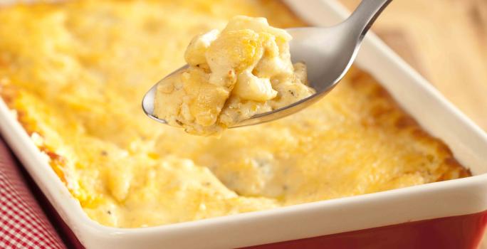 fotografia em tons de marrom, vermelho, amarelo e branco tirada de uma tábua de madeira, por cima um recipiente retangular vermelho e branco com a batata gratinada dentro.