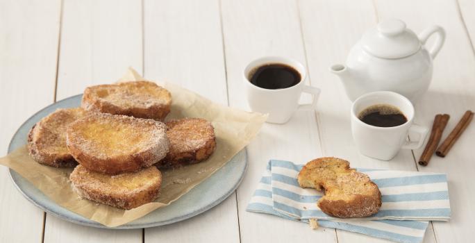 Fotografia em tons de dourado e branco, com rabanadas fritas sobre papel manteiga e prato azul, ao lado serviço de café, paus de canela e rabanada sobre guardanapo listrado em azul e branco, tudo sobre bancada em tom pastel.