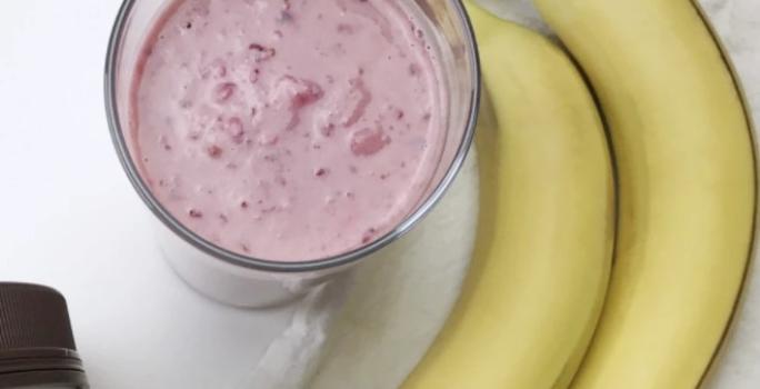 Fotografia em tons de rosa em uma mesa branca, uma toalha branca, duas bananas e um copo de vidro alto com o smoothie de morango com banana e pasta de amendoim dentro dele.