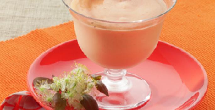 Fotografia em tons de laranja em uma mesa de madeira com uma toalha laranja, um pano vermelho desenhado com flores, um prato vermelho e uma taça de vidro com a mousse de pitanga dentro e uma pitanga em cima para decorar.