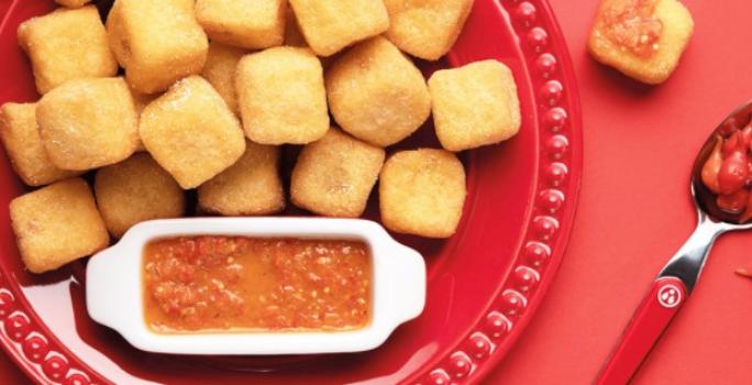 Fotografia em tons de vermelho em uma mesa vermelha, com um prato vermelho raso grande e vários dadinhos de polenta recheados com queijo coalho. Ao lado, um potinho com o molho de pimenta biquinho.