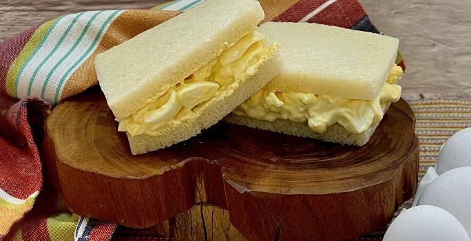Foto da receita de Sanduíche de Ovos. Observa-se uma tábua de madeira com dois sanduíches de pão de forma sem casca com o recheio de ovos dentro sobre a tábua.
