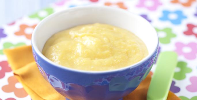 Fotografia em tons de azul em uma bancada de madeira azul clara, um pano com flores coloridas, um paninho laranja, um recipiente azul com a papinha dentro dele. Ao lado, uma colher infantil verde.