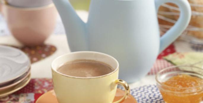 foto em tons de marrom, bege e azul tirada de um pratinho bege com uma xicara bege que contém o chocolate quente dentro. Ao fundo um bule azul