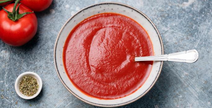 fotografia em tons de cinza e vermelho de uma bancada cinza vista de cima. Ao centro um recipiente redondo com sopa de tomate e um colher para servir, e ao lado esquerdo contém tomates.