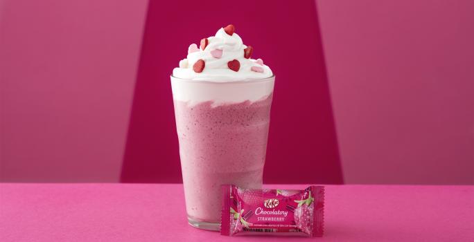 Foto da receita de Frappé. Observa-se um fundo rosa com um copo alto decorado de chantily e confeitos de coração com um kit kat de morango na frente.