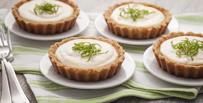 Fotografia em tons de verde em uma mesa de madeira com um pano listrado em verde e quatro pratinhos brancos cada um com a torta creme de limão. Ao lado, três garfos.