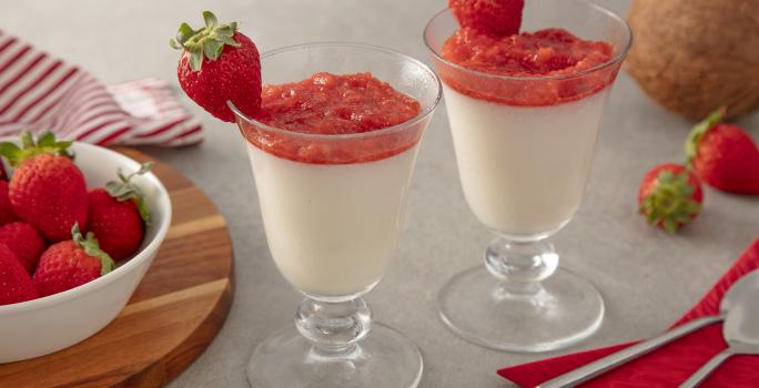 Foto da Receita de Taças de coco com purê de morango. Observa-se duas taças de vidro com o mousse e o purê com morangos decorando a borda da taça. Como decoração, vemos morangos ao lado direito em um potinho e um coco fresco ao lado direito