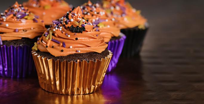Foto de cupcakes cobertos por um creme laranja, decorados com confeitos coloridos em embalagens brilhantes nos tons de roxo e laranja.