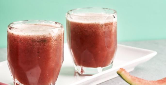 Fotografia em tons de rosa e vermelho em uma bancada de madeira clara com um recipiente branco retangular com o suco de melancia e menta. Ao lado, fatias de melancia e uma melancia inteira.
