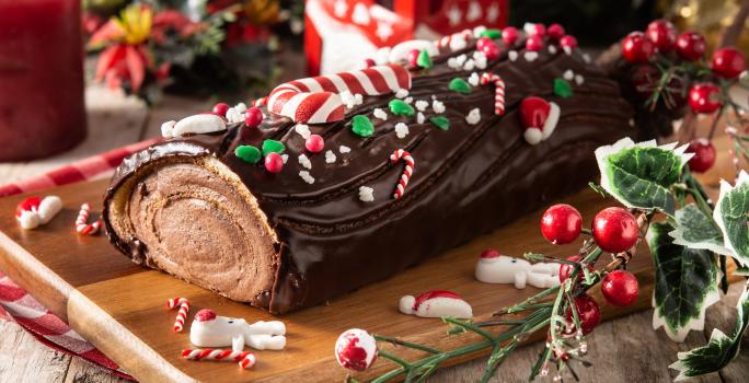 fotografia em tons de marrom, verde e vermelho tirada de uma tábua de madeira. Por cima contém um rocambole de chocolate em formate de troco de arvore com enfeites de Natal.