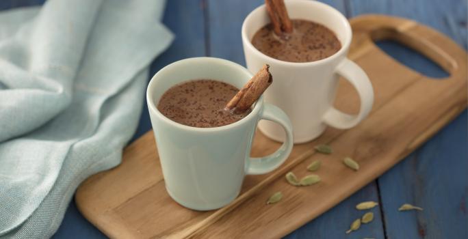 Fotografia em tons de azul em uma bancada de madeira azul, um pano azul, uma tábua de madeira com duas xícaras brancas com o chocolate quente com canela dentro delas.