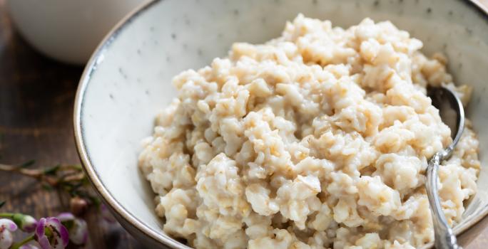 Fotografia em tons claros de um recipiente de vidro branco com acaçá cremoso, e no mesmo recipiente, uma colher de sopa apoiada dentro do acaçá. Ao fundo, molho em um recipiente pequeno na cor branca, sobre uma mesa de madeira na cor marrom escuro.