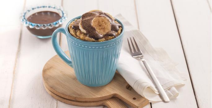 Foto da receita de bolo de caneca com cacau e banana em uma caneca azul, sobre tábuas de madeira, com um guardanapo de pano branco e um garfo sobre ele.