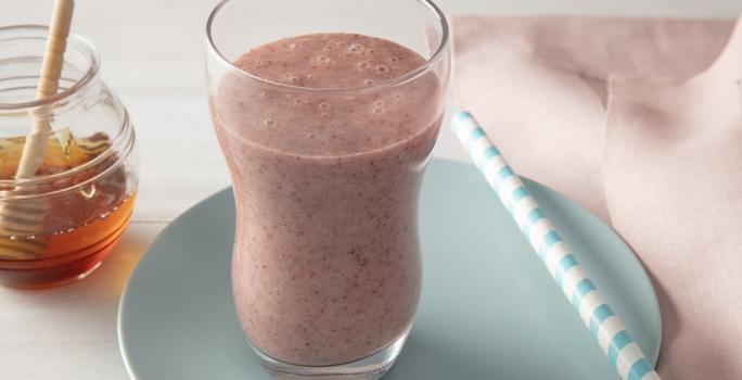 Foto da receita de smoothie iogurte molico com morango e chia servida em um copo largo sobre um pratinho azul com um canudo listrado ao lado junto deum paninho rosa. Ao fundo há um vidro de mel