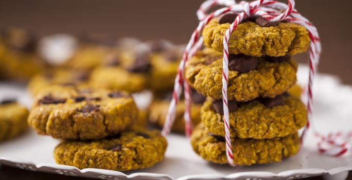 Fotografia em tons de amarelo em uma bancada de madeira com um recipiente branco raso e vários cookies integrais de aveia e abóbora em cima dele.