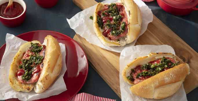 Foto em tons de vermelho da receita de choripan tradicional servida em três porções, duas sobre uma tábua de madeira e uma sobre um prato vermelho. Ao fundo há dois bowls de porcelana vermelhos além de uma panelinha e um paninho vermelhos