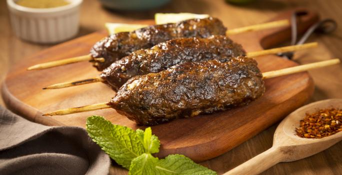 Fotografia em tons de marrom, verde e cinza de uma bancada de madeira marrom vista de frente. Uma tábua de madeira contém 3 espetinhos com carne moída, ao lado uma colher de madeira com tempero, um pano cinza e um potinho redondo branco com tempero.