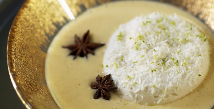 Fotografia de ovos nevados com um creme inglês, algumas raspas de limão por cima e dois anis estrelados no creme. A sobremesa está dentro de um recipiente fundo de alumínio na cor dourado, que está sobre uma mesa preta.