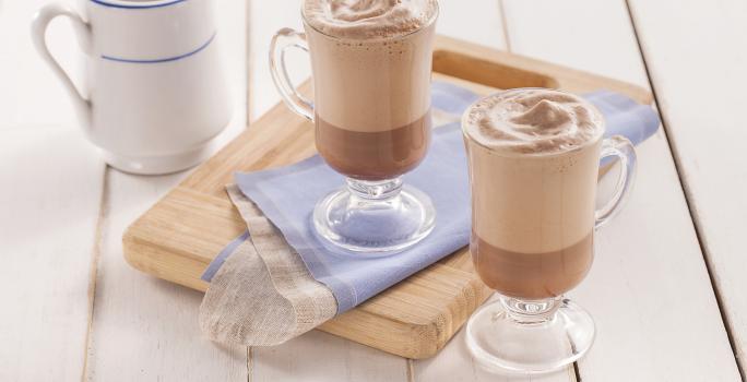 Fotografia em tons de azul em uma bancada de madeira clara, uma tábua de madeira, um paninho azul e duas xícaras de vidro com o cappuccino gelado de cacau dentro deles. Ao fundo, uma jarra branca com azul com mais cappuccino.