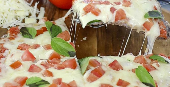 Foto aproximada de uma pizza marguerita, decorada com tomates e folhas de manjericão, sobre uma tábua de madeira e com tomates ao fundo