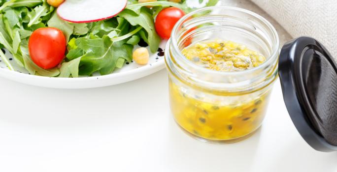 fotografia em tons de branco, verde e amarelo de uma bancada branca vista de cima. Contém um pote transparente com molho de maracujá e ao lado um prato redondo branco com salada.