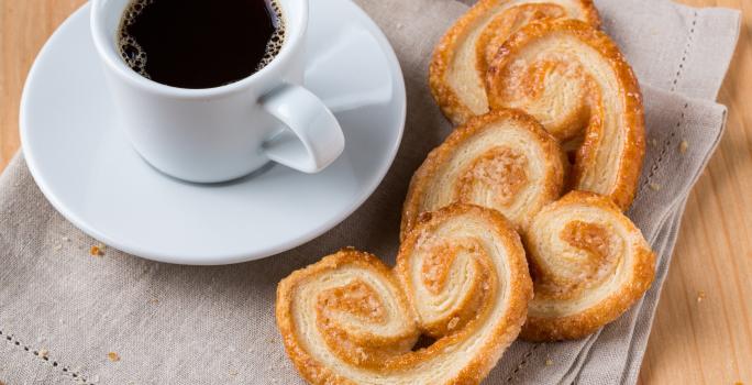 Foto da receita de Palmier ou Orelha de Macaco. Observa-se o biscoito em disposto em cima de um pano de prato e, ao lado, uma xícara com café sobre um pires.