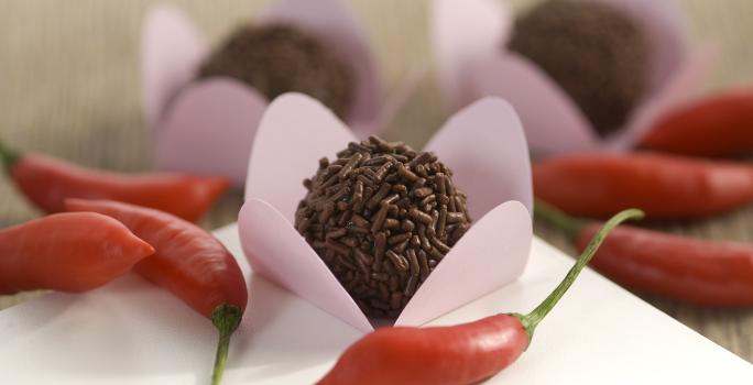 Fotografia em tons de vermelho em uma bancada de madeira com uma toalha branca, ao centro um brigadeiro em uma forminha rosa e várias pimentas vermelhas espalhadas pela mesa.