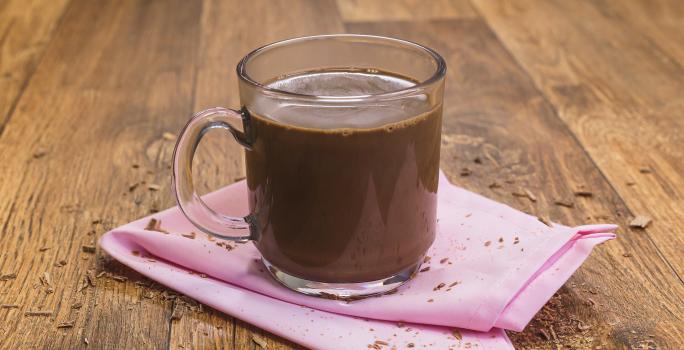 Fotografia de uma bancada de madeira com um paninho rosa e uma xícara transparente com o chocolate quente dentro.