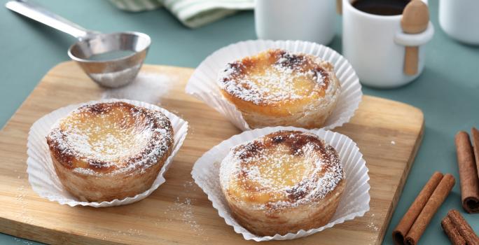 Fotografia em tons de amarelo em uma bancada de madeira de cor azul. Ao centro, uma tábua de madeira contendo os pasteis. Ao lado, há uma peneira de inox e ao fundo, duas xícaras de café.