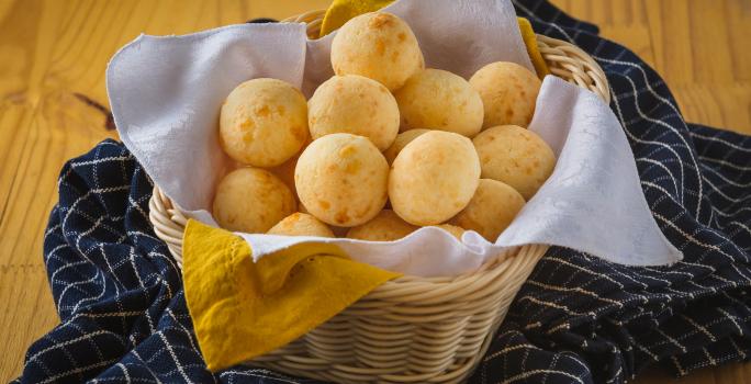 Foto aproximada de uma cesta de vime com diversos pães de queijo dentro. Ela está sobre uma bancada de madeira com um tecido azul quadriculado.