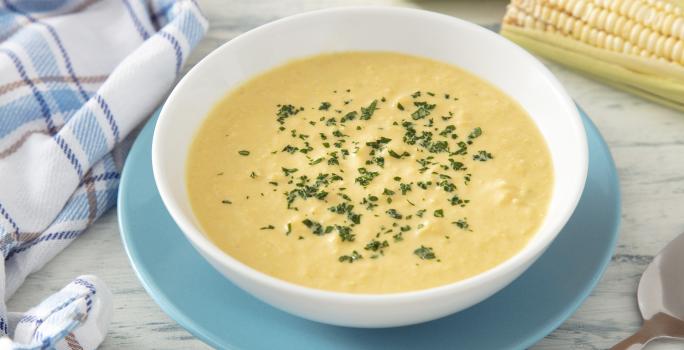 Fotografia em tons de amarelo e azul em uma bancada de madeira clara, um prato azul e branco ao centro com sopa de milho. Ao lado uma espiga de milho e um paninho azul quadriculado.