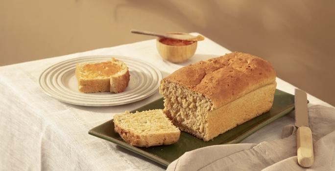 Foto em tons de bege da receita de pão com farinha de aveia servida sobre uma base de porcelana verde-escuro cortada a uma fatia com uma faca de cozinha ao lado. Ao fundo há outra fatia do pão servida em um prato bege claro e um pote de madeira com mel