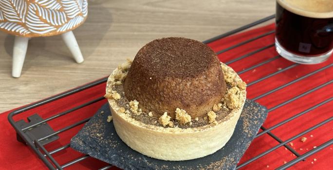 Fotografia de uma mini tortinha com base de biscoito maisena com um pequeno bolo de chocolate ao topo. Ao fundo existe um pequeno copo com café e embaixo uma tolha de cor vermelho escuro