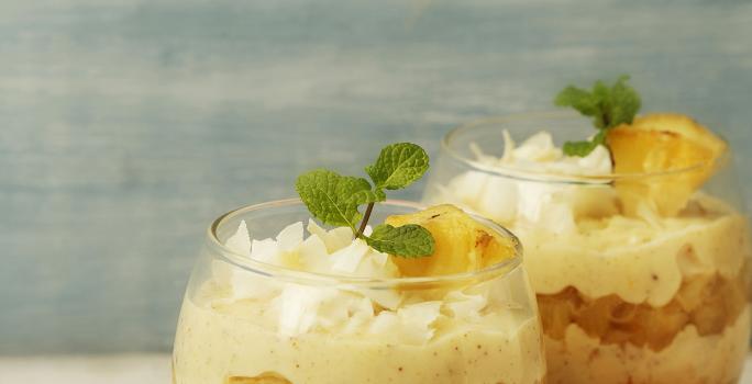 Fotografia em tons de laranja em uma bancada de madeira azul, um pano colorido listrado, duas taças de vidro com o doce de abacaxi com creme de Leite Moça dentro delas.