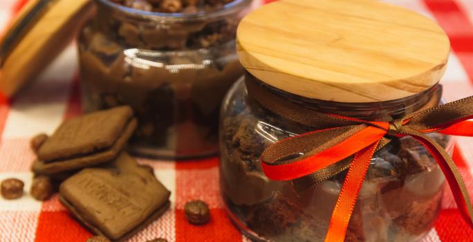 Foto de uma bancada forrada com uma toalha quadriculada vermelha e branca. Sobre ela há 2 potes de vidros com a receita de Brownie de Nescau com Brigadeiro. Um deles está fechado e o outro aberto. Além disso há 2 biscoitos e algumas bolinhas de cereal