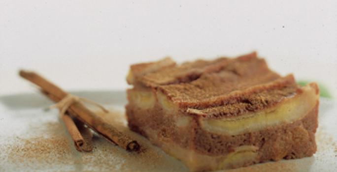 Fotografia em tons de canela e cinza, com pedaço de torta de banana com canela sobre travessa verde com ramos de canela ao lado, tudo polvilhado com canela, em fundo cinza.