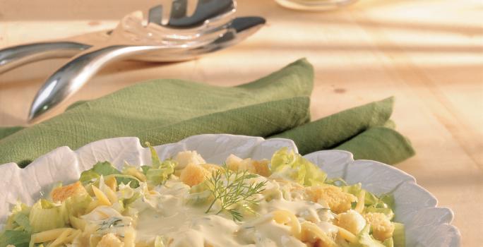 Fotografia em tons de verde em uma bancada de madeira clara, um pano verde, um pegador de salada e um prato redondo branco fundo com a salada de alface, queijo, torradinhas e iogurte dentro. Fotografia em tons de verde em uma bancada de madeira clara, um pano verde, um pegador de salada e um prato redondo branco fundo com a salada de alface, queijo, torradinhas e iogurte dentro.