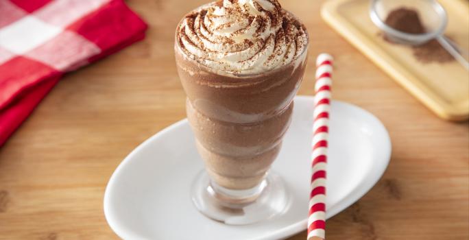 Foto da receita de Milkshake de Cappuccino. Observa-se um copo alto com a bebida decorado com chantilly e polvilhado de chocolate em pó. Ao lado direito, um canudo vermelho e branco para decorar.