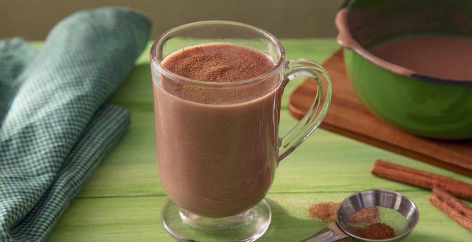 Fotografia em tons de marrom em uma bancada de madeira de cor verde. Ao centro, uma xícara contendo o chocolate quente. Ao lado, há uma toalha verde quadriculada e ao fundo, uma tábua de madeira com uma panela verde em cima.