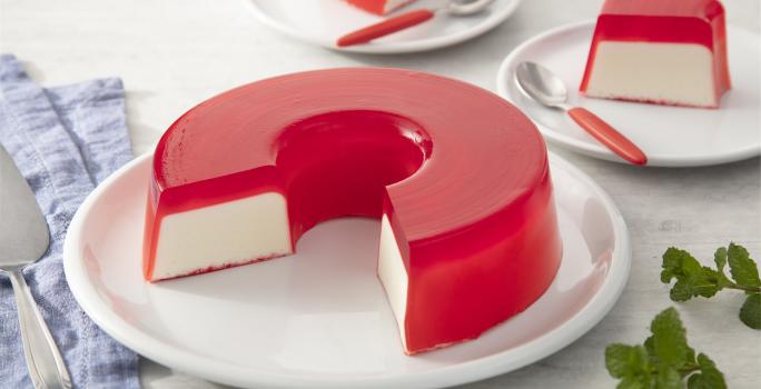 Fotografia em tons de rosa em uma bancada de madeira clara, um paninho azul, um prato branco redondo raso com a gelatina de morango recheada com creme branco. Ao fundo, pratinhos brancos com fatias da gelatina e talheres com cabo vermelho.