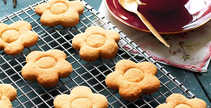 fotografia em tons de azul e bege de uma bancada azul vista de cima, contém uma grade com biscoitinhos em formato de flores e ao lado um pratinho vermelho com uma xicara vermelha que contém a bebida de chocolate branco.