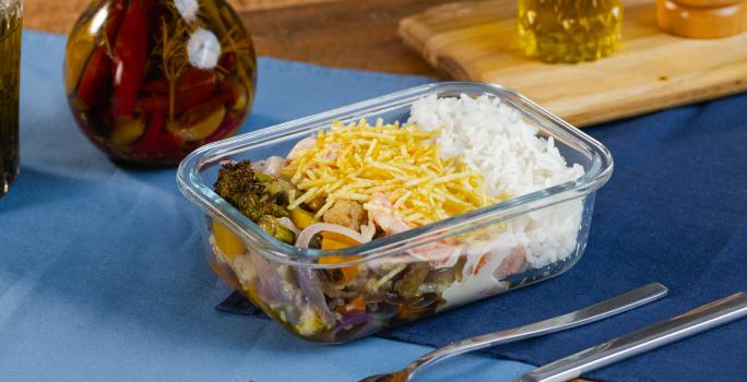 Fotografia de uma marmita retangular pequena com arroz, fricassê, batata palha, arroz e legumes assados. A marmita está sobre dois panos sobrepostos de cor azul, ao lado de talheres e uma mini tábua com pimenta e azeite.