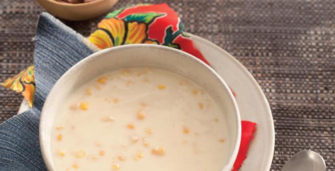Fotografia em tons de cinza e  branco de um paninho cinza, sobre ele um prato redondo branco raso  com um paninho estampado, sobre ele um prato redondo fundo branco com a canjica. Ao lado uma faca com cabo de madeira. Ao fundo dois potinhos com