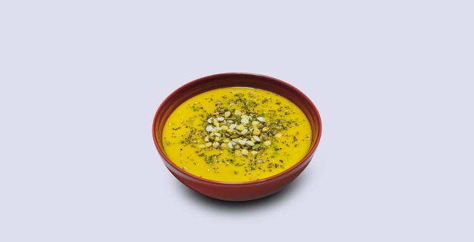 Fotografia em tons de amarelo em um fundo branco com um prato fundo vermelho e o caldo de abóbora e carne seca com Maggi dentro.