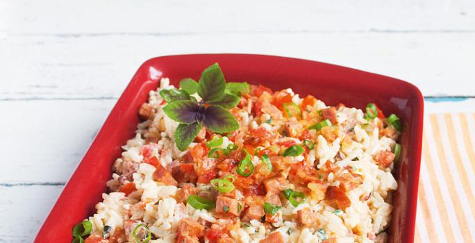 Fotografia em tons de vermelho e branco de uma bancada branca com uma pano listrado laranja e branco, uma travessa vermelha retangular com o arroz, pedaços de tomate e linguiça e manjericão.