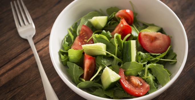 Fotografia de um recipiente fundo branco com uma salada de folhas, pepino e tomates. Ao lado do recipiente, um garfo, que está sobre uma mesa de madeira escura.