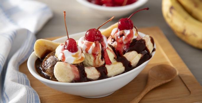 Fotografia em tons de vermelho em uma bancada de madeira de cor cinza. Ao centro, uma tábua de madeira com uma tigela em cima contendo o banana split. Ao lado, há uma colher de madeira e um pano listrado azul. Ao fundo, 2 bananas e algumas cerejas.